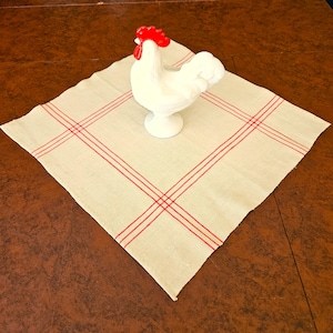 May include: A white ceramic rooster figurine with a red comb sits on a cream-colored linen napkin with red plaid stripes. The napkin is placed on a brown wooden surface. The rooster is a decorative item.
