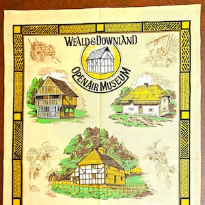 May include: A cream-colored tea towel featuring black and yellow illustrations of historic buildings and the text Weald & Downland Open Air Museum. The design includes a central logo, a cart, and various timber-framed houses with thatched roofs.