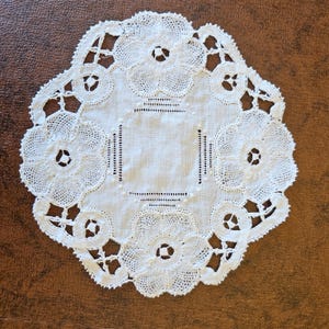 May include: A white, round doily with an intricate floral lace design. The doily features six large flower motifs and a central square, all connected by delicate openwork. The edges are scalloped, adding to its vintage charm.