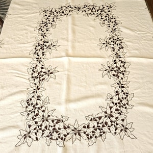 May include: A rectangular off-white tablecloth featuring a central border of dark brown embroidered poinsettia flowers and leaves. The embroidery forms a continuous oval-shaped frame pattern that repeats along the edges of the fabric surface.