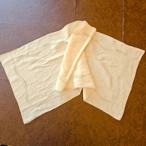 May include: A rectangular off-white table runner featuring a subtle, delicate embroidered border pattern in a matching light thread. The fabric has a soft, woven texture with clean, straight edges and a folded section in the center.