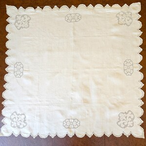 May include: A square off-white tablecloth featuring a scalloped edge with intricate white embroidery. The fabric displays four corner motifs of swirling floral patterns and four side motifs of symmetrical flower designs in white thread.
