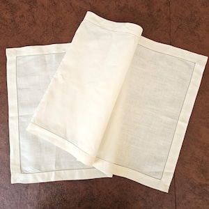 May include: Off-white linen table runner with a dark gray stitched border. The runner is partially rolled, revealing its texture and design. The fabric appears to be lightweight and suitable for dining or decorative use.