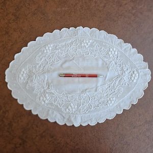 May include: Oval white embroidered doily with a scalloped edge, featuring a grape and leaf design. A red pen with the word "AVIATOR" is placed on the doily. The doily is on a brown surface.
