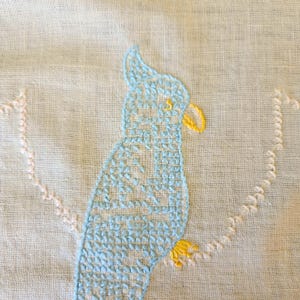 May include: Embroidered light blue parrot with yellow beak and feet on a light background. The bird is stitched with a cross-stitch pattern. The design is part of a larger textile piece, possibly a pillow or wall hanging.