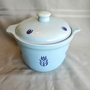May include: A light blue ceramic casserole dish with a matching lid and two side handles. The exterior of the pot and the lid are decorated with a repeating dark blue tulip flower pattern. The lid features a central knob handle for easy removal.