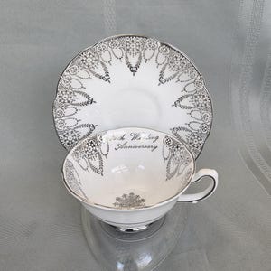 May include: White teacup and saucer set with silver filigree detailing. The teacup and saucer have a scalloped edge. The teacup has the words "25th Wedding Anniversary" printed on it.