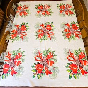 May include: A cream-colored tablecloth with a repeating pattern of red bows, pinecones, and green foliage. The bows have a bright red color, and the pinecones are brown. The green foliage includes pine needles and holly leaves. The tablecloth is spread out on a wooden table.