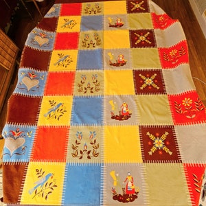 May include: A colourful patchwork quilt with embroidered squares. Designs include birds, flowers, and figures. The quilt features red, yellow, blue, brown, and grey squares, with white stitching throughout.