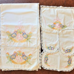 May include: A set of off-white fabric linens featuring embroidered floral fan motifs in pink, yellow, and blue. The pieces are finished with delicate white crocheted lace edging along the borders, showcasing intricate needlework and floral patterns.