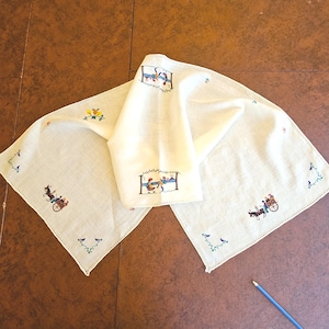 May include: A cream-colored linen napkin featuring delicate multi-colored embroidery of figures in horse-drawn carts, floral motifs, and small birds. The fabric has a hemstitched border and is displayed with a blue pencil for scale reference.
