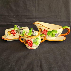 May include: A cream-colored ceramic serving set featuring a butter dish, sugar bowl, and gravy boat. Each piece is decorated with a grape cluster design in red, purple, and green, with gold trim. The set is designed for serving food.