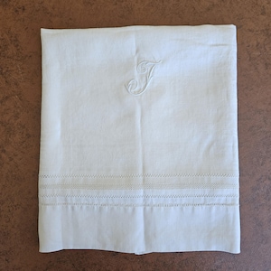 May include: White linen towel with an embroidered letter "J" in the upper right corner. The towel features a decorative stitched border near the bottom. The background is a solid brown color.