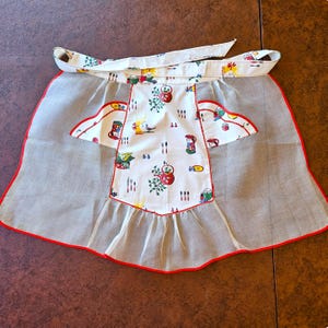 May include: A white half apron featuring a central panel with a colorful kitchen-themed print of red teapots, cutlery, and birds. The apron has sheer gray side panels, two pockets with red scalloped trim, and a matching white waistband with ties.