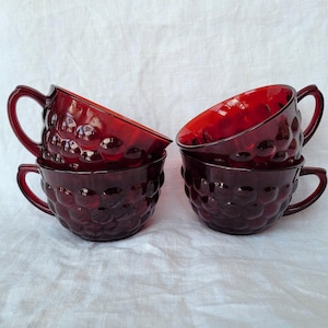 May include: Four ruby red glass teacups stacked on a white surface. The cups have a textured, bubble-like pattern and curved handles. The cups are angled, showcasing their interior.