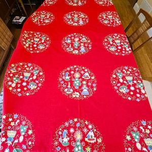 May include: A red Christmas tablecloth with circular designs featuring festive illustrations. Each circle contains images of Christmas trees, houses, and figures, surrounded by floral and snowflake patterns. The tablecloth is spread across a wooden table.