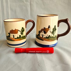 May include: Two ceramic mugs with a cream-colored base, brown handles, and a blue stripe. Each mug features a painted landscape scene of a house and trees. A red marker with the text "universal" is also present.
