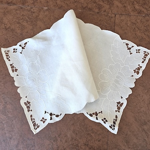 May include: A folded, off-white, scalloped-edge doily with floral embroidery. The doily features a large flower design with detailed leaves and cut-out accents along the edge. The fabric appears to be linen or a similar material.