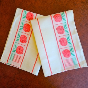 May include: Two off-white kitchen towels featuring a repeating vertical pattern of red apples with green leaves. Each towel includes thin red stripes running vertically along the fabric, with a decorative horizontal line detail at the bottom edge.