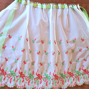 May include: A white apron with red and green embroidered bows and sprigs. The apron has a scalloped edge and green ribbon ties. The top edge has green ribbon loops and a red trim.