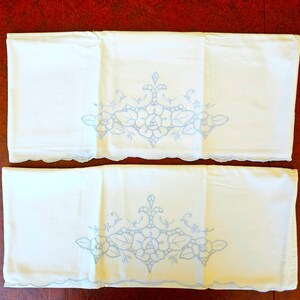 May include: Two white pillowcases with a light blue floral embroidery design. The embroidery features roses and leaves, with a scalloped edge. The pillowcases are folded and displayed on a dark wooden surface.
