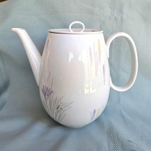 May include: A white porcelain teapot featuring a delicate floral pattern with light purple and yellow crocus-like flowers. The teapot has a curved handle, a tapered spout, and a circular lid with a small loop handle on top for easy removal.