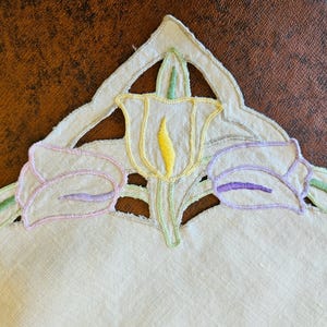 May include: A beige fabric corner featuring intricate cutwork and embroidery of three tulips. The central tulip is yellow with green stems, while the two side tulips are outlined in pink and purple thread, creating a decorative floral border.