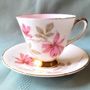 May include: A vintage teacup and saucer set. The white porcelain cup and saucer feature a pink floral design with gold trim. The inside of the cup is pink. The cup has a gold handle and sits on a gold base.