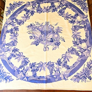 May include: A square tablecloth with a blue and white design. The border features a repeating pattern of grapes and leaves. The center of the tablecloth has a still life of fruits, including grapes, pears, and a pineapple.