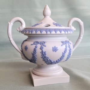 May include: A pale lavender ceramic urn with a matching lid, featuring light blue relief floral swags and ribbon motifs. The vessel has two curved handles, a pointed finial on the lid, and rests on a square base, showcasing intricate detailing.