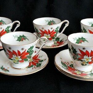 May include: A set of six white teacups and saucers. Each piece is decorated with red poinsettias, green holly, and gold detailing. The teacups have curved handles and sit on matching saucers, ideal for serving tea during the festive season.