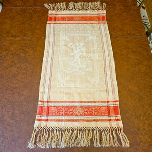 May include: Cream-colored rectangular textile with red and blue decorative borders and fringe. The center features a white detailed design of a figure with floral elements. The textile is likely a decorative table runner or wall hanging.