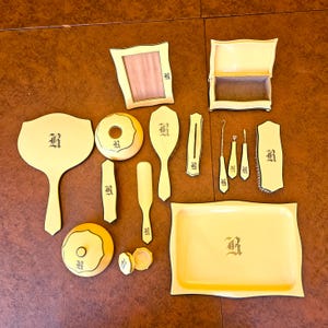 May include: A collection of antique vanity items in a cream color with black trim. The set includes a hand mirror, brush, tray, and other accessories, each bearing an ornate letter "R". The items are arranged on a brown surface.