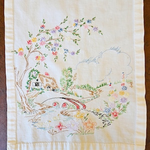 May include: Embroidered linen table runner featuring a pastoral scene. The design includes a thatched-roof cottage, a bridge over a stream, and colorful flowers. The embroidery uses various colors, including red, green, and blue.