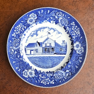 May include: A blue and white decorative plate featuring a detailed illustration of a building with a flag, surrounded by floral patterns. The plate's center displays the text "Birthplace of Calvin Coolidge, Plymouth Vermont."