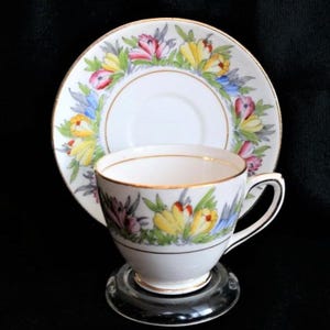 May include: A white porcelain teacup and matching saucer set featuring a hand-painted floral pattern of yellow, pink, and blue crocus flowers with green leaves. Both pieces are finished with a thin gold trim along the rims and base.