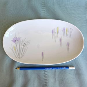May include: White oval serving platter with a floral design. The platter features purple, yellow, and grey stylized flowers and foliage. A blue pencil is placed next to the platter for scale.