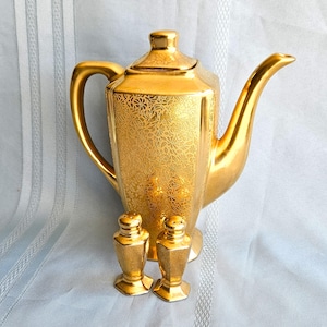May include: A gold-colored tea set with a floral pattern, including a teapot and two salt shakers. The teapot has a curved spout and handle. The salt shakers are small and hexagonal. The set is displayed on a light blue surface.