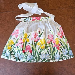 May include: A white apron with a floral design featuring pink, yellow, and red irises, along with blue hydrangeas and green leaves. The apron has a scalloped edge and white ties.