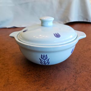 May include: A light blue ceramic bowl with a matching lid and two side handles. The bowl and lid are decorated with a repeating pattern of small, dark blue tulip illustrations. The piece has a smooth, glossy finish and a rounded, circular shape.