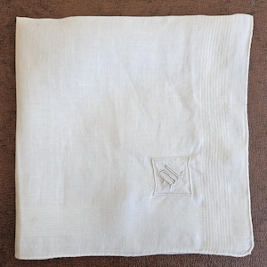 May include: A square white linen handkerchief featuring a subtle woven border pattern and a small, embroidered monogram in the lower right corner. The fabric has a visible weave texture and clean, straight edges throughout the design.