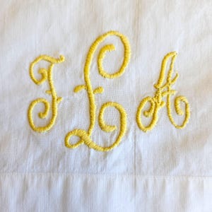 May include: White fabric with yellow embroidered initials F, L, and A. The letters are in a cursive script, with a decorative, flowing design. The embroidery is centered on the fabric.