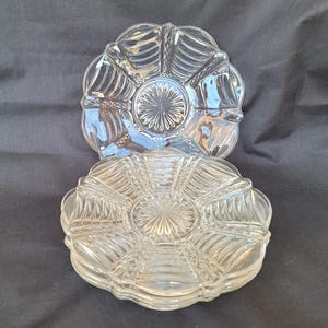 May include: Two clear glass serving dishes. Each dish has a scalloped edge and is divided into sections radiating from a central, starburst-like design. The dishes are stacked, with one partially visible behind the other, on a dark background.
