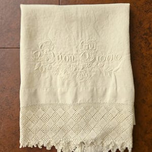 May include: Cream-colored linen towel with embroidered floral details and the words "Buon Giorno". The bottom edge features a decorative lace trim with a scalloped edge. The towel is folded and displayed on a brown surface.