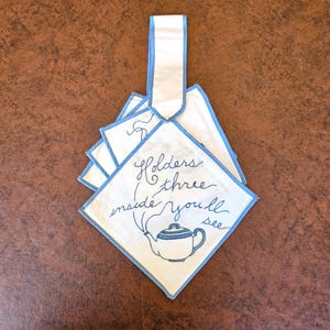 May include: White square handkerchiefs with blue trim and a blue teapot illustration. The handkerchiefs have the handwritten text "Holders three inside you'll see". The handkerchiefs are stacked and connected by a white ribbon with blue trim.