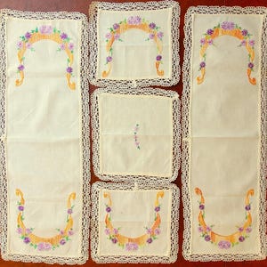 May include: A set of five cream-colored linens featuring intricate crochet lace borders. Two long rectangular runners and three square napkins are decorated with embroidered orange arches and purple floral accents along the edges.