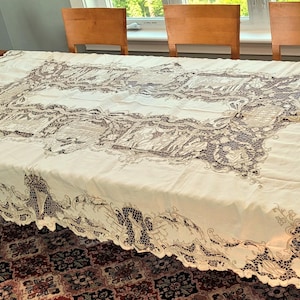 May include: Cream-colored tablecloth with intricate lace detailing. The rectangular cloth is draped over a wooden table, showcasing a detailed floral and geometric lace pattern. The edges of the cloth are scalloped, adding a decorative touch.