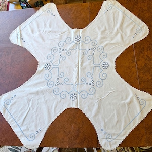 May include: White, four-pointed tablecloth with blue embroidered floral and swirl designs. The edges are scalloped and trimmed with blue stitching. The tablecloth is laid out on a brown surface.