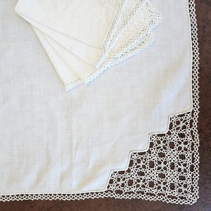 May include: White linen tablecloth with a scalloped edge and intricate lace detailing. Several matching napkins are neatly stacked in the upper left corner. The tablecloth and napkins are a bright white color.