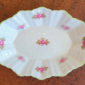 May include: An oval-shaped white ceramic dish with a scalloped edge trimmed in light green. The surface features a repeating pattern of small pink rose blossoms with green leaves arranged around the center and along the fluted rim.